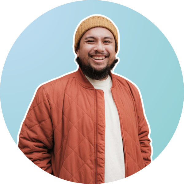 A smiling man with a beard wears a mustard beanie, white shirt, and orange quilted jacket, standing against a light blue gradient background framed in a circular border, perfect for a social media scheduler | plannthat.com